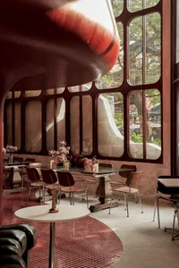 Modern restaurant interior with red walls, large windows, and natural light, featuring tables, chairs, and potted plants