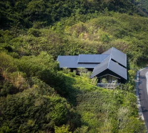 A house with a black roof sits on a hillside surrounded by trees and greenery in a serene natural environment