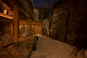 Outdoor dining area at night with tables, chairs, and potted plants in front of a lit building on a gravel pathway with a stone wall