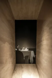A dining area with a table set for two, featuring white linens, silverware, wine glasses, and a vase with flowers