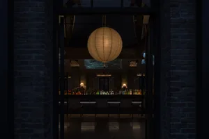 A cozy restaurant with a round lamp as a centerpiece, surrounded by chairs and dining tables, with bottles on surfaces throughout