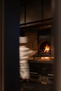 A man in a chef's uniform stands near an open fireplace in a kitchen with a brick wall, bottles on surfaces, and a cozy atmosphere