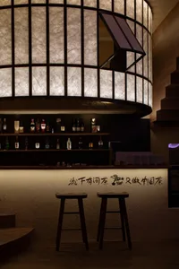 A cozy Asian restaurant bar area with a wooden counter and stools, featuring numerous bottles and an inviting atmosphere