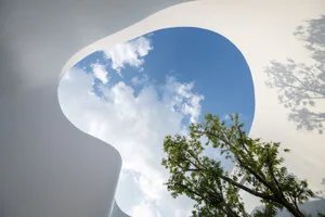 An open space with a large white wall featuring a round hole, allowing sunlight to enter and illuminate the area, surrounded by trees outside