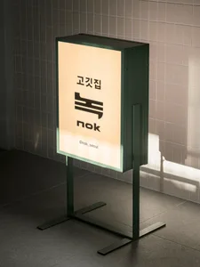 An illuminated advertisement sign on a green stand in an urban setting, possibly in Asia