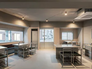 An empty kitchen with white walls and floors, featuring two dining tables, several chairs, and three sinks, appearing clean and well-organized