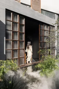 A woman stands in front of a doorway to a dark room with a wooden door frame, two chairs, and a potted plant