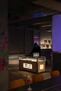 Office space with a wooden table, glass display case, and modern furniture, featuring a desk with a laptop and two people