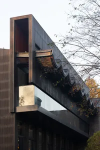 A modern building with a black facade featuring a large window with a metal frame, two smaller windows below, and a possible concrete or similar material construction, receiving ample natural light