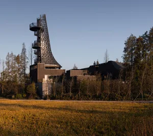 A modern building with a ship's bow design, situated in a rural area surrounded by trees and grassy fields, blending harmoniously with nature
