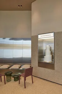 A wedding cake on a silver tray in a glass case, surrounded by chairs and potted plants in a hotel lobby-like setting