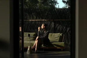 A woman sits on a couch in a calm and relaxing indoor living space with a dining table, chairs, and a vase in the background, near a window with natural light