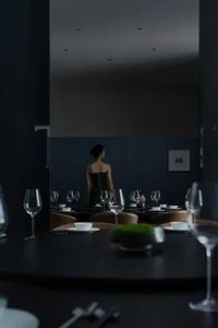 A woman stands in front of a dining table set with wine glasses, dinnerware, and a mirror, possibly in a restaurant or private event space
