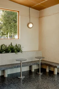 A modern dining space with concrete walls, featuring a bench and two dining tables, surrounded by potted plants, creating a calm and inviting atmosphere