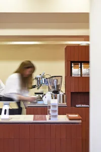 A barista prepares drinks at a busy coffee shop counter with various items and customers in the background