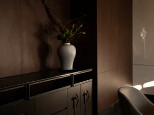 Dimly lit room with a white vase on a table, containing flowers, and a chair against the wall, with two drawers nearby
