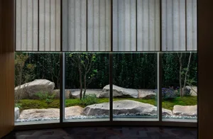 A tranquil indoor space with a large window offering a view of a garden with a pond, rocks, and plants, surrounded by trees, and natural light pouring in