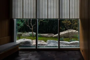 A cozy indoor space with a large window, rock wall, potted plants, and vases, offering a view of a garden outside