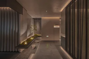 A modern hotel lobby with white walls, gray flooring, and two potted plants, one near the left side and one near the center, with a bench at the right edge