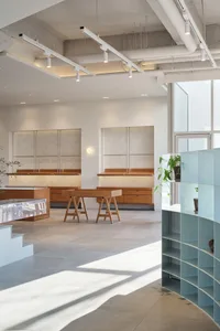 Open living area with white walls, wooden floors, and natural light from a large window, featuring chairs and potted plants