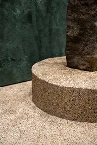 A stone building with a modern design featuring a large stone column in front of a green wall, with two other columns nearby, showcasing a combination of gray and green tones