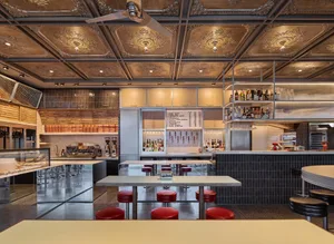A restaurant bar with wooden elements, multiple chairs, and tables, featuring a ceiling fan and various items on the tables