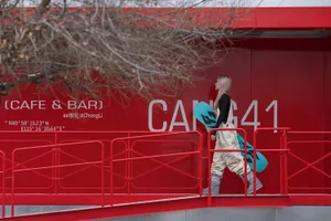 Two people in a casual indoor setting with a red wall featuring white lettering and a metal staircase with a railing