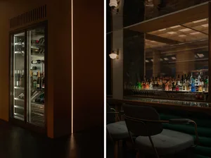 A modern bar area with a green wall, refrigerator, chairs, and bottles, with a couch and dining table in the background