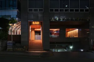 A modern Asian restaurant at night with a brick exterior, glass windows, and a warm interior atmosphere, featuring dining tables, chairs, and bottles