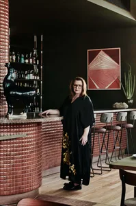 A woman poses at a bar counter with bottles and chairs, surrounded by a casual and social atmosphere