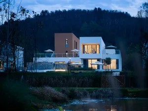 A serene and inviting modern house at night with a white exterior, brown accents, multiple windows, doors, and a balcony, set against a dark background