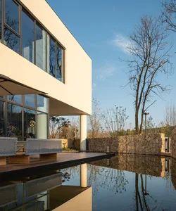 Modern house with a large glass wall overlooking a pond, featuring a living area with couches and chairs, and an exterior made of concrete