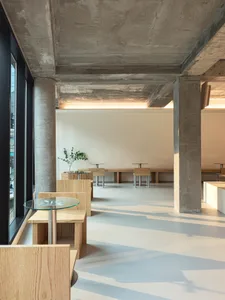 Architectural rendering of a large open space with concrete floors and walls, featuring tables, chairs, and potted plants, designed for socializing or gatherings