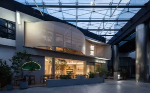 A modern Asian restaurant with a glass building, curved roof, white walls, and wooden accents, featuring dining tables and chairs, and potted plants