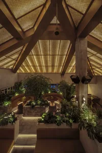 A modern restaurant interior with high ceilings, wooden beams, and stone elements, featuring potted plants and dining tables in a welcoming atmosphere