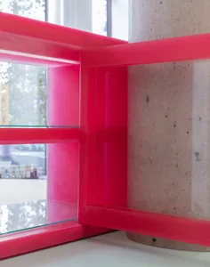 A modern display case with a red interior and glass walls stands in front of a window, allowing natural light to enter the area