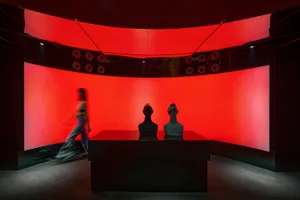 A modern gallery space with red walls, black tables, and a display of three sculptures on a central table