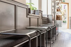 Modern restaurant interior with a long dining table and metal chairs, surrounded by potted plants and natural light
