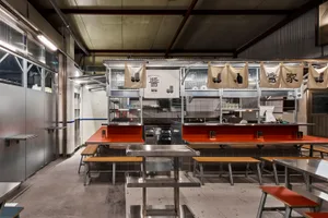 Industrial kitchen with two dining tables, chairs, and scattered bottles, featuring a functional and practical atmosphere with a focus on efficiency and organization