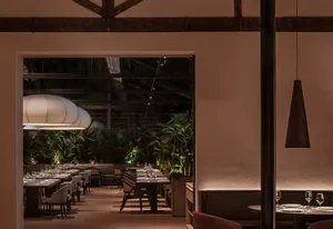 Dimly lit dining area with a large wooden dining table, multiple chairs, and potted plants, suggesting a high-end restaurant or hotel interior