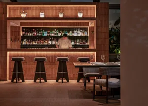 A cozy bar area with wooden walls and floors, featuring a bar counter and chairs, with a man tending to customers