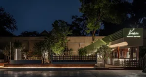 A serene restaurant at night with a green roof, white walls, and lush greenery surrounding the building, including potted plants near the entrance and on the sides