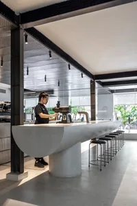 一間現代咖啡廳，設有長形吧台與高腳椅，周圍環繞座椅，並有大型窗戶帶來自然光線，一位女性站在吧台後方