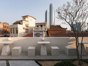 Outdoor cafe scene with two skyscrapers in the background, tables with white tops, chairs, and potted plants on a patio area