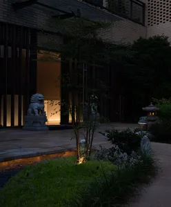 A serene Asian-style courtyard with statues, decorative elements, and a blend of traditional and modern architecture, illuminated by natural and artificial light, surrounded by plants and trees