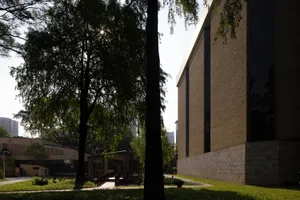 A modern building with a brick facade, surrounded by trees, with sunlight illuminating the scene, creating shadows and highlights on the building's surface