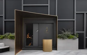 A restaurant entrance with a black door, gold trim, and a dining area with a table and chairs, surrounded by a potted plant
