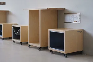 Three wooden cabinets on wheels in front of a white wall, each with a unique design, and a small sign above one of the cabinets