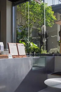 A modern coffee shop with a glass wall, natural light, and potted plants, featuring a counter with cups, bowls, and a vase