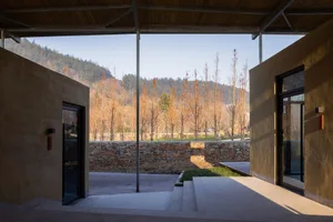 A rustic building facade with stone pillars and a patio area with a bench, surrounded by trees and a calm atmosphere